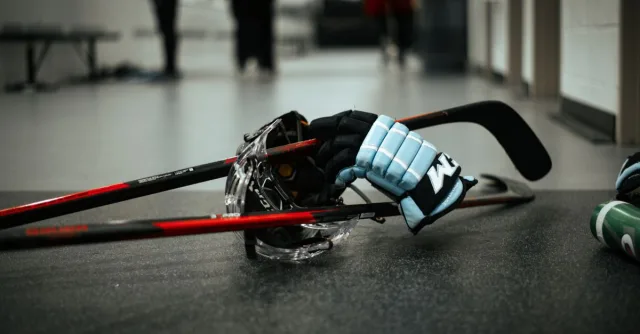 Close-up of hockey gloves, helmet, and sticks on an indoor ice rink floor, conveying sports readiness.