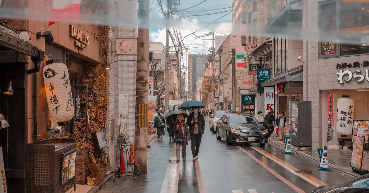 A lively street in Japan with pedestrians and shops under rainy weather.