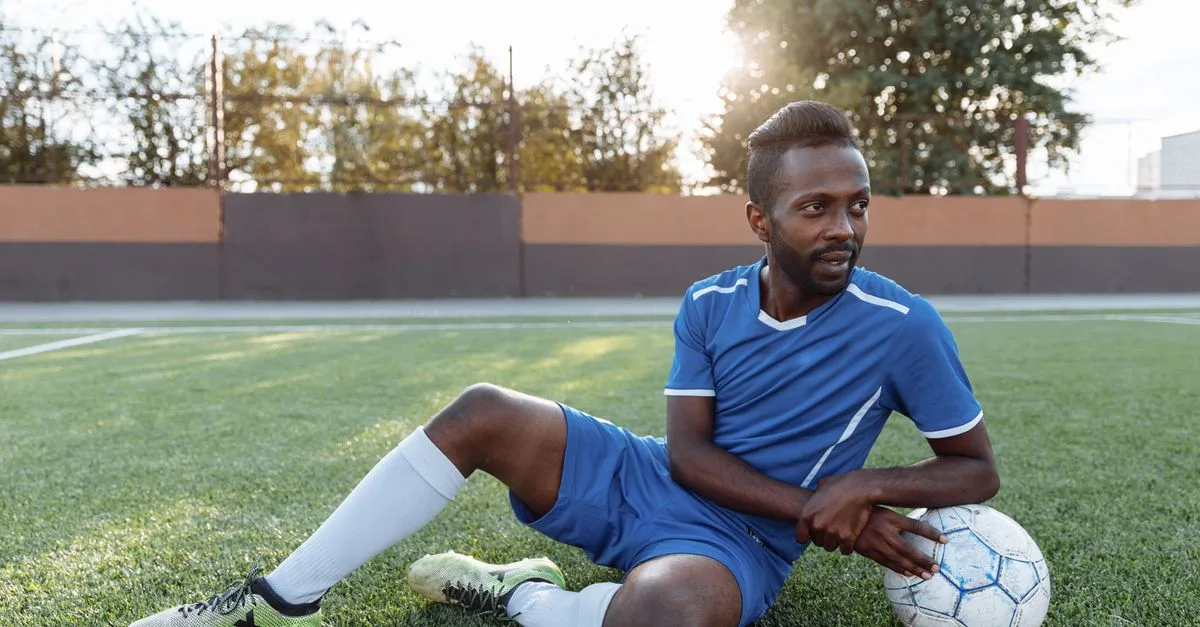 A soccer player in blue jersey sitting on a green field holding a ball, enjoying a sunny day.