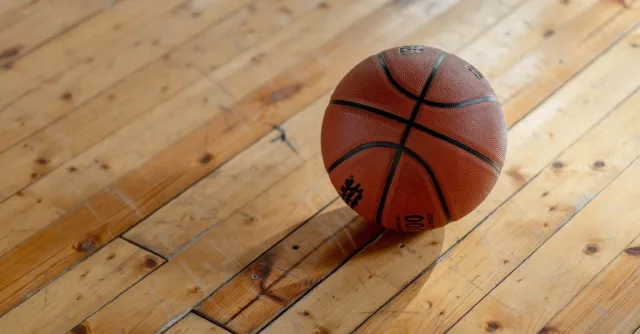 A single basketball resting on a polished wooden gym floor, casting a shadow.