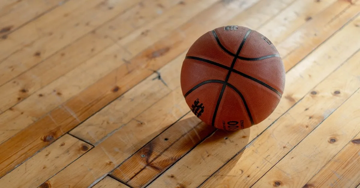 A single basketball resting on a polished wooden gym floor, casting a shadow.