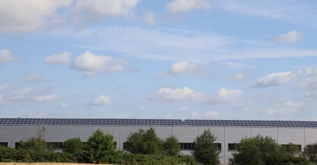 A countryside warehouse with solar panels and fields under a blue sky with clouds.