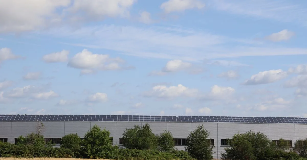 A countryside warehouse with solar panels and fields under a blue sky with clouds.