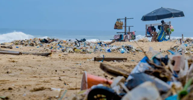 Sandy beach cluttered with trash and ocean debris, with vendors under an umbrella nearby.