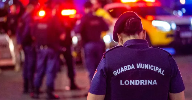 A municipal guard stands on duty in the vibrant streets of Londrina at night.