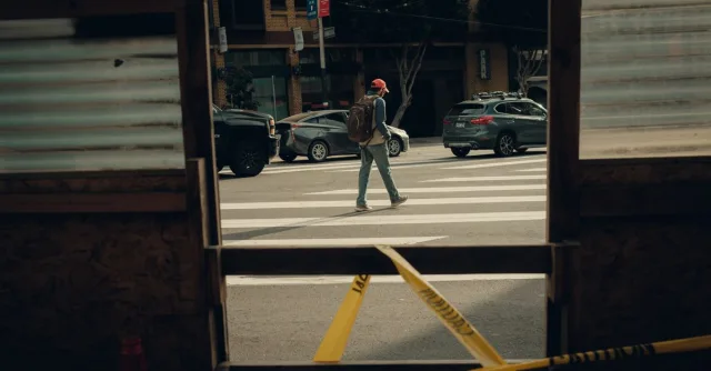 A man with a backpack crosses a city street through a cordoned off construction area.