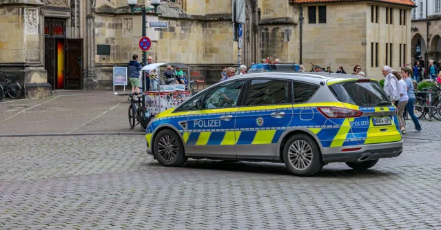 A police car stationed in a historic square of Münster, Germany, surrounded by pedestrians and bicycles.