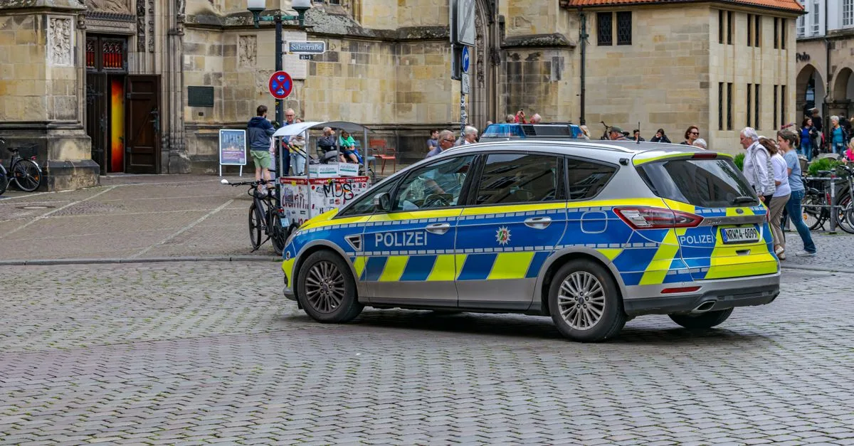 A police car stationed in a historic square of Münster, Germany, surrounded by pedestrians and bicycles.