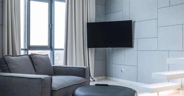Interior of living room with gray sofa near table on carpet and windows with curtains near TV and white stairs