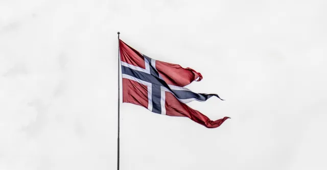 Norwegian national flag waving on a flagpole against a cloudy sky, symbolizing national pride.