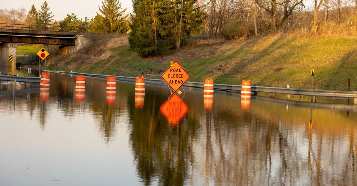 A flooded road with warning signs indicating 'Road Closed Ahead' due to water overflow.
