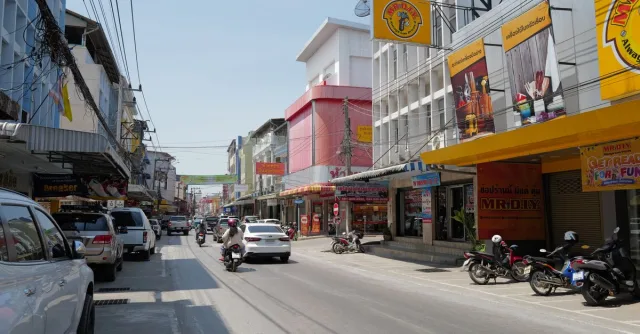 Lively Thai city street scene with cars, motorcycles, and colorful shop facades on a sunny day.