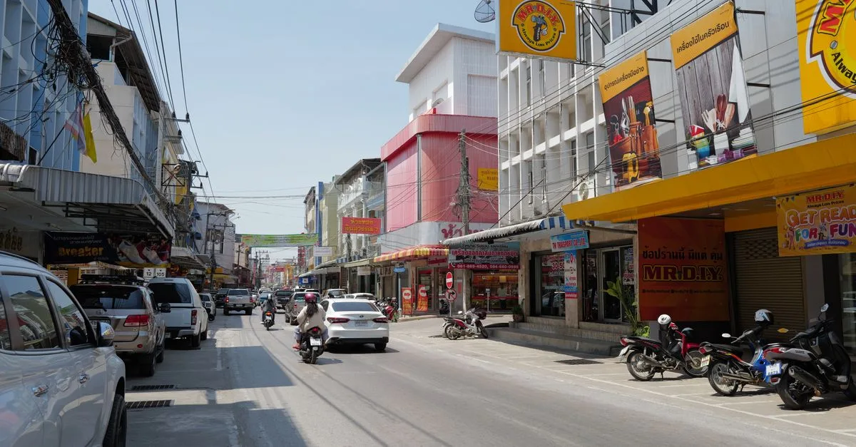 Lively Thai city street scene with cars, motorcycles, and colorful shop facades on a sunny day.