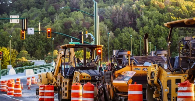 Heavy machinery and barriers at a road construction site with traffic lights and forest backdrop.