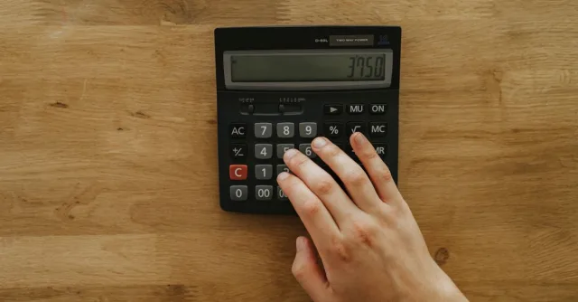 A person's hand pressing keys on a calculator displaying 3750 on a wooden table, top view.