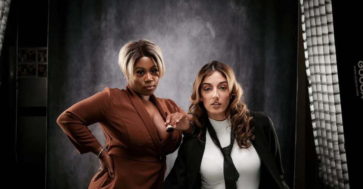 Two women pose confidently in professional attire in a studio setting.