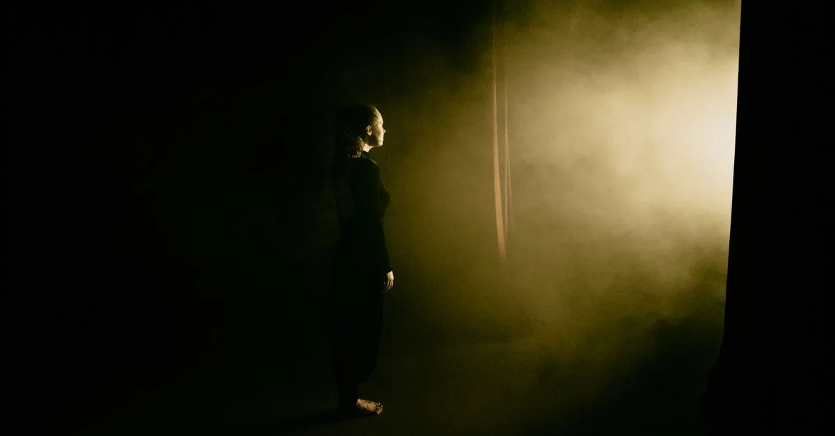 Side view of a woman standing on a smoky stage, illuminated dramatically from the side.