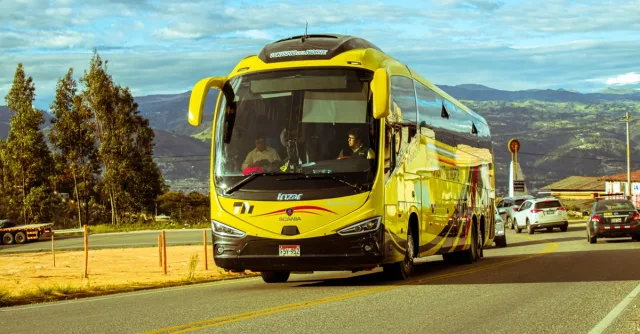 A bright yellow bus travels on a scenic mountain road under a blue sky, surrounded by cars and nature.