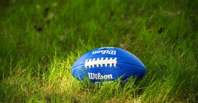 A bright blue Wilson football resting on a lush green grass field, illuminated by sunlight.