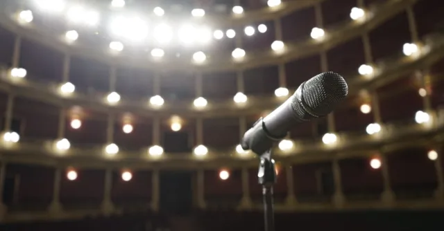 Close-up of a microphone on stage with bright theater lights in the background.