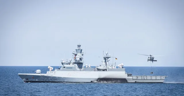 A naval frigate navigates open waters with a hovering helicopter, showcasing maritime operations.