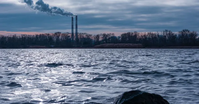 Factory smoke stacks emitting pollution over river with dramatic sky in Kyiv.