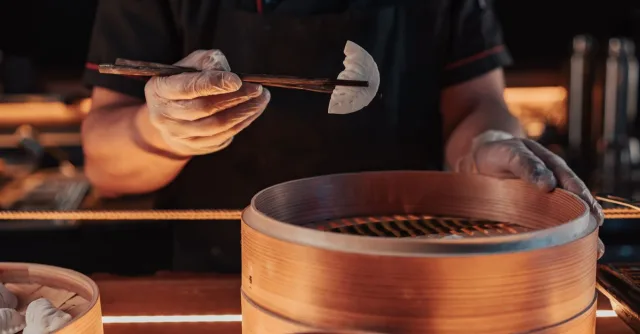 Chef using chopsticks to place dumplings in a bamboo steamer for cooking.
