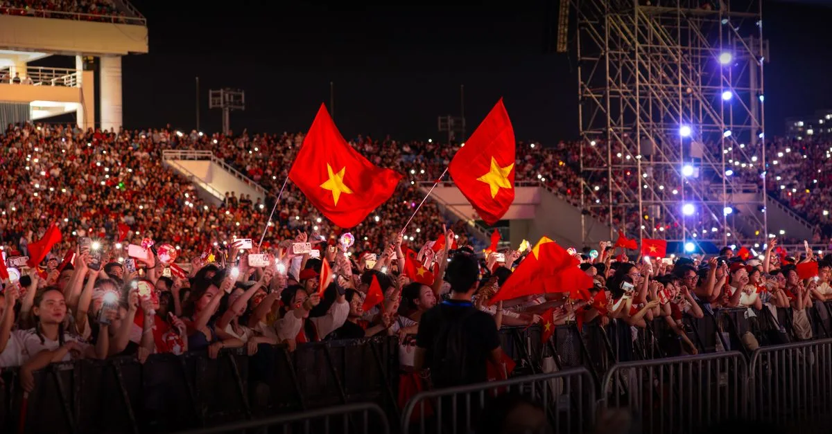 A nighttime concert with a large, energetic crowd waving flags under bright lights.
