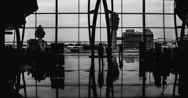 Black and white image of travelers silhouetted against large airport windows, capturing urban travel ambiance.