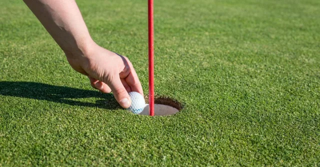 A hand retrieves a golf ball from the hole on a well-manicured green, signifying a successful putt.