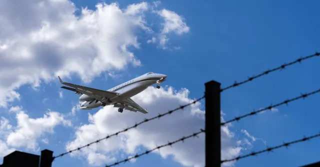 Jet approaching for landing, silhouetted against a bright blue sky with clouds and barbed wire in foreground.