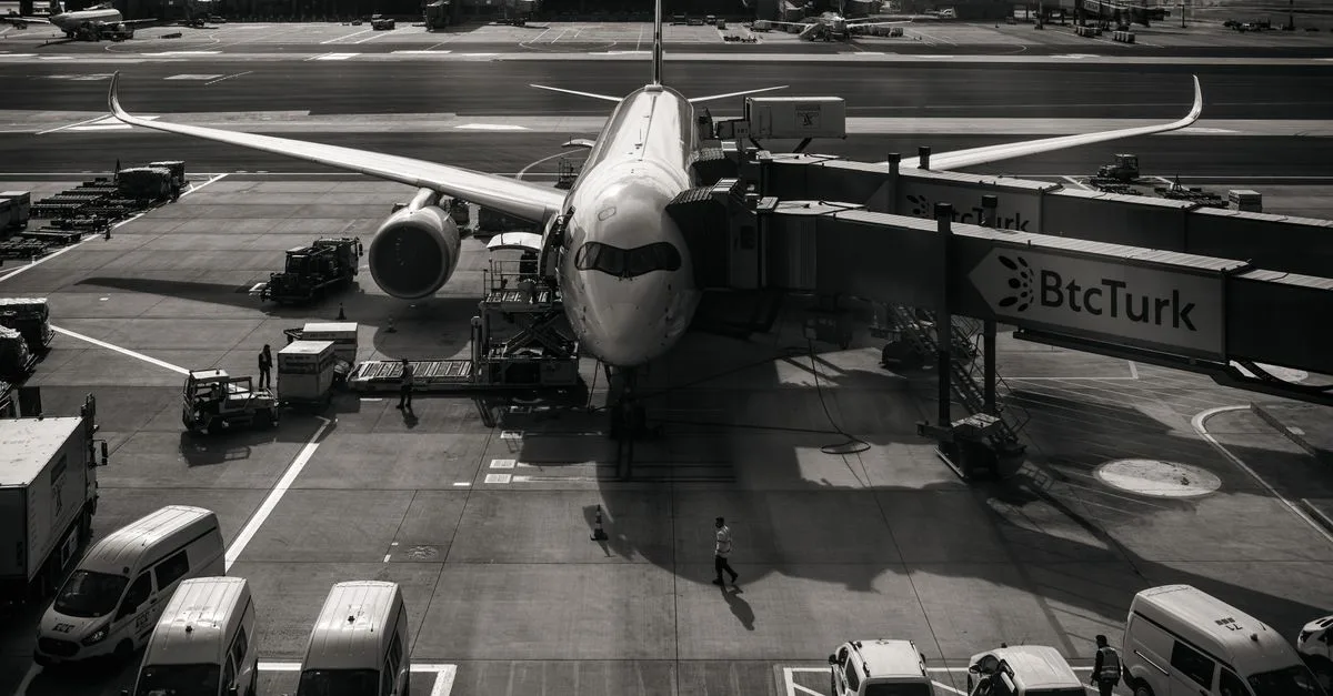 Monochrome image of airplane at airport gate with vehicles and jetway during the day.
