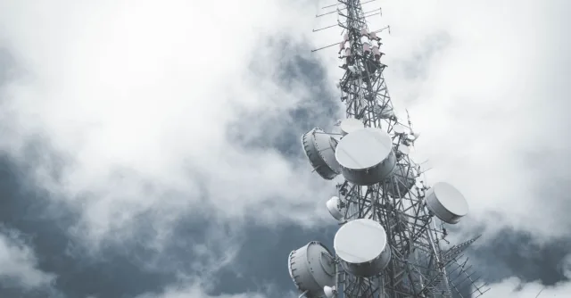 A towering communication mast with satellite dishes, set against a cloudy sky.