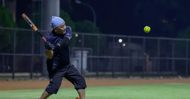 Male player batting during a nighttime softball game under stadium lights.