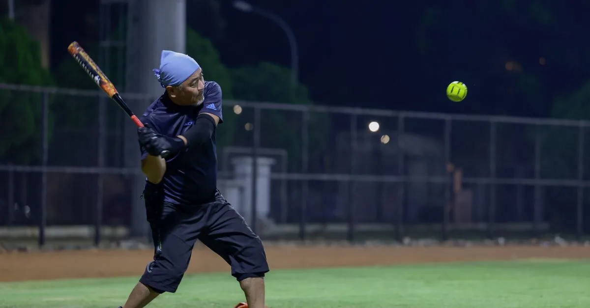 Male player batting during a nighttime softball game under stadium lights.
