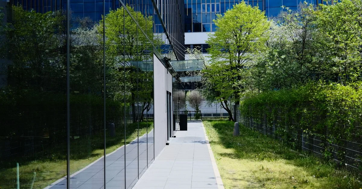 Modern glass building with pedestrian walkway and greenery, reflecting urban architecture.
