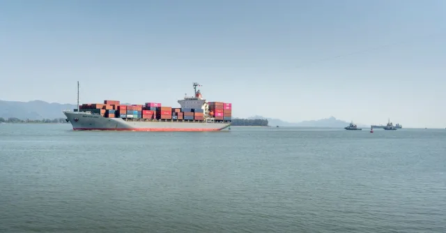 A large cargo ship travels across the open ocean, framed against distant mountains under a clear sky.