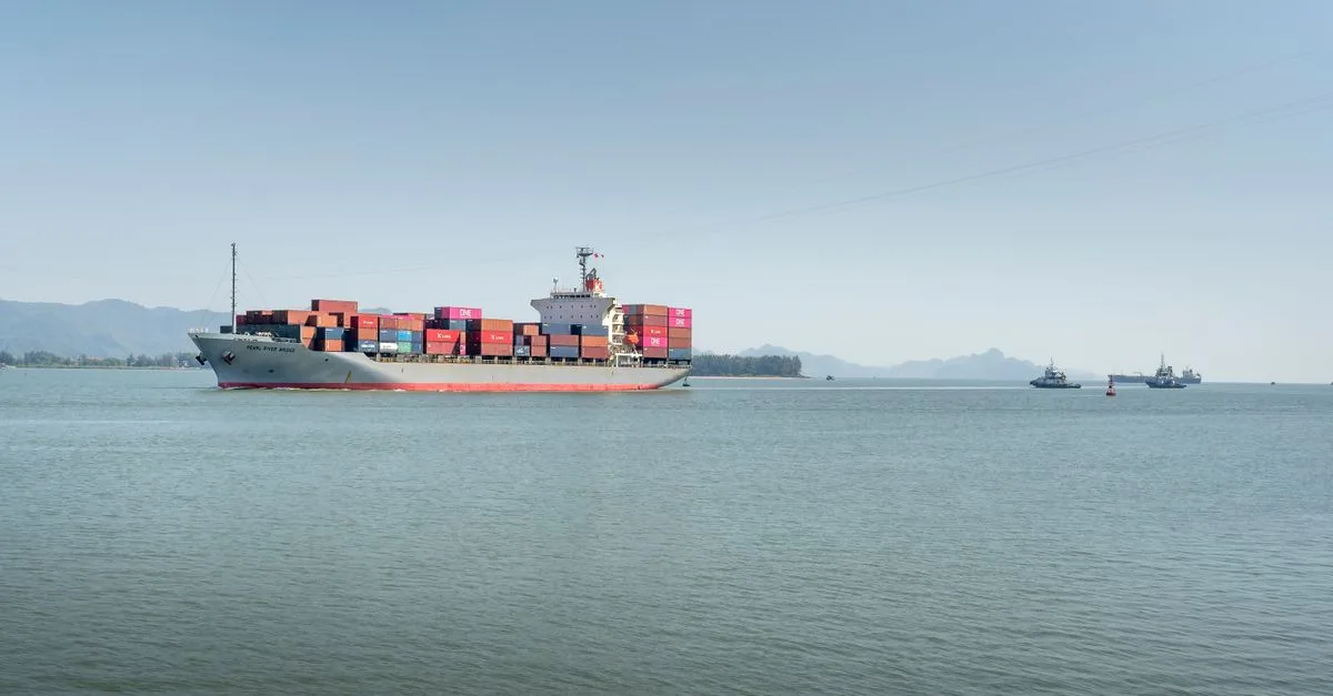 A large cargo ship travels across the open ocean, framed against distant mountains under a clear sky.