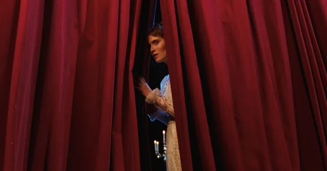 Woman peeks through red theater curtains, capturing a theatrical moment.