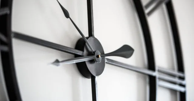 Stylish black round shaped clock with hands and Roman numeral hanging on white wall in light room on blurred background