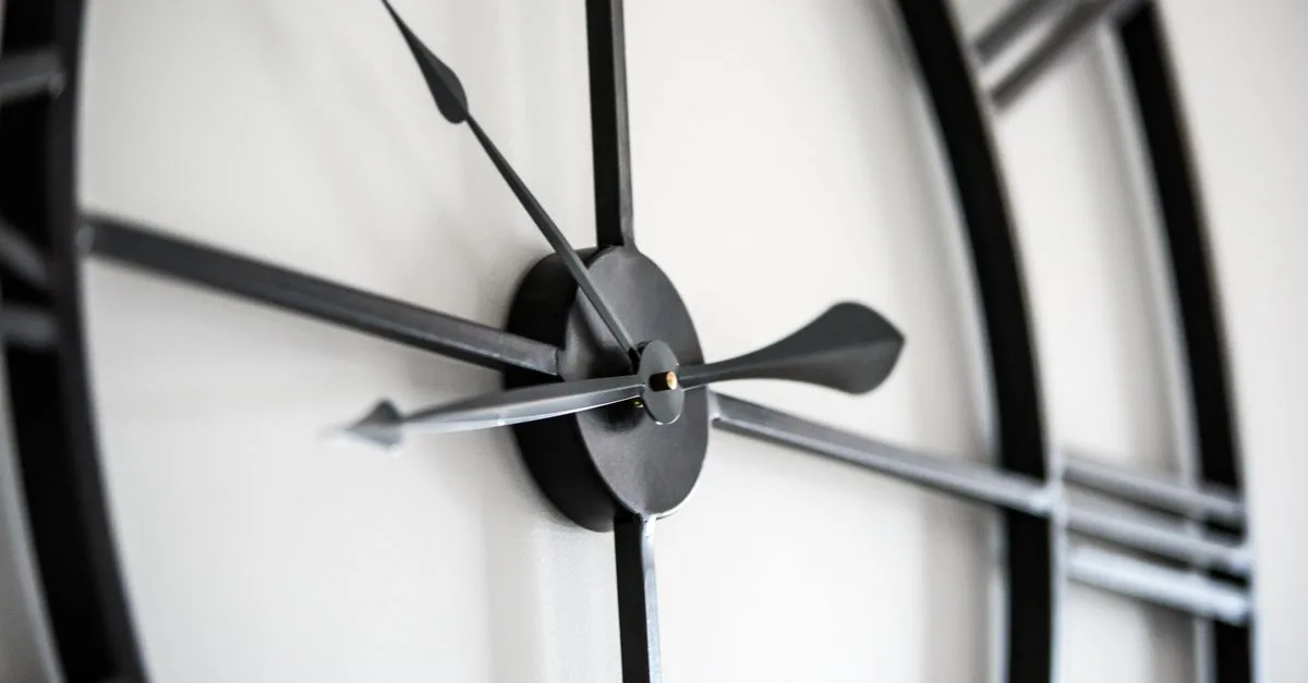Stylish black round shaped clock with hands and Roman numeral hanging on white wall in light room on blurred background