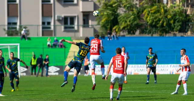 Soccer players in red and blue uniforms compete fiercely during an outdoor match on a sunny day.