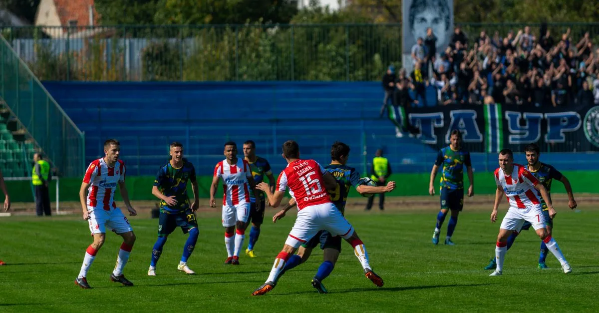 Intense soccer game with teams in action under bright sunlight, surrounded by cheering fans.