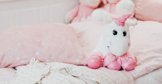 A soft unicorn plush toy sits on a pastel pink bed with pillows and blankets in a child's room.