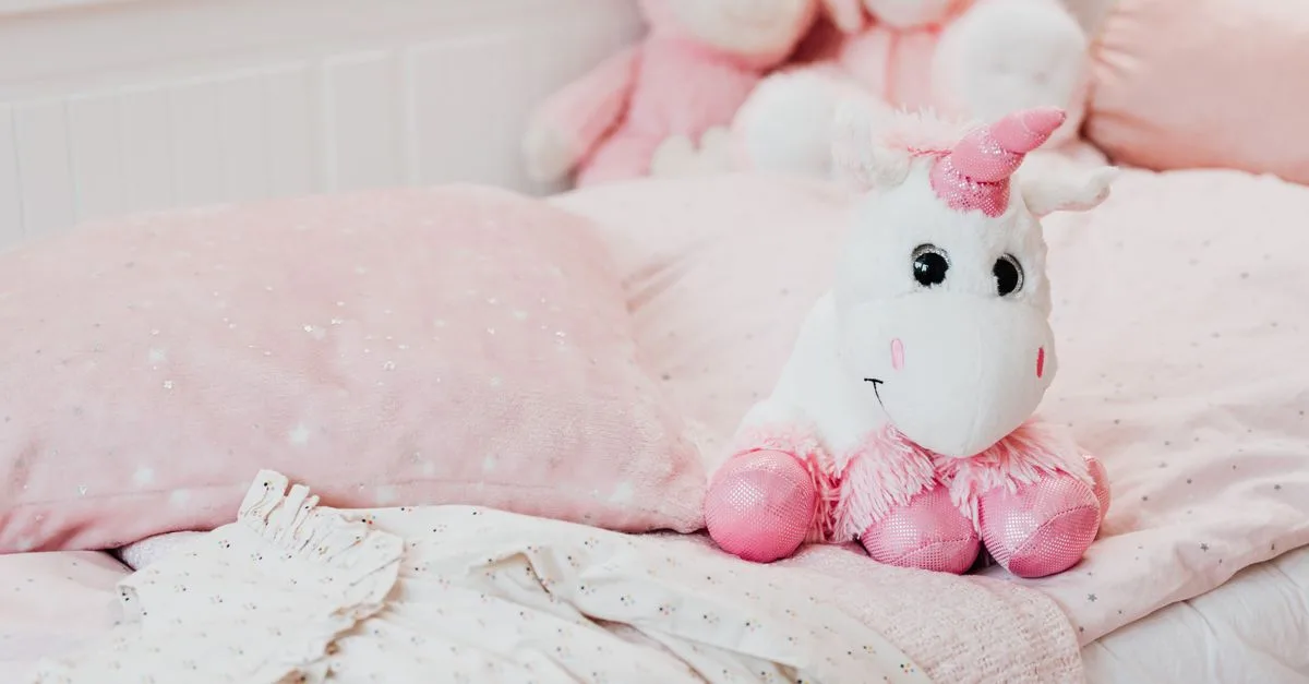 A soft unicorn plush toy sits on a pastel pink bed with pillows and blankets in a child's room.