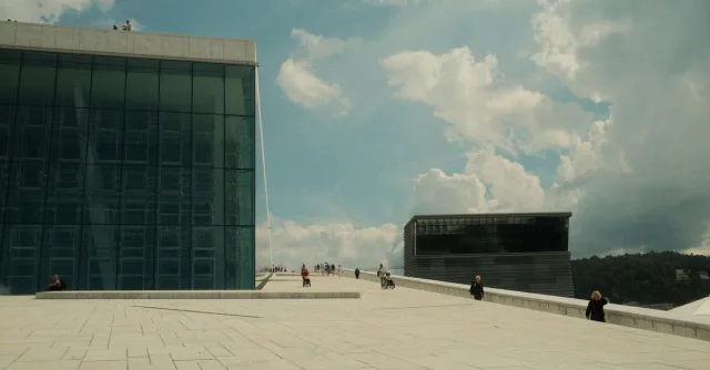 Wide angle view of Oslo Opera House with people on its sloped roof under a cloudy sky.
