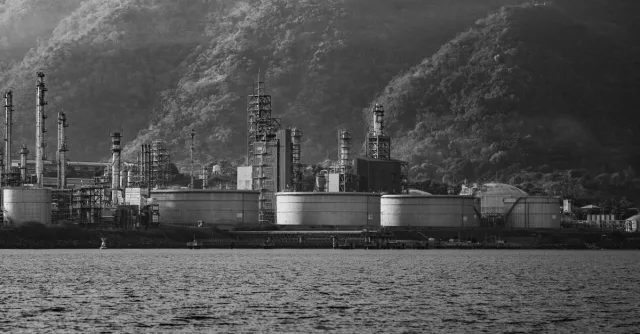 Black and white image of an industrial facility by a mountain range and a body of water.