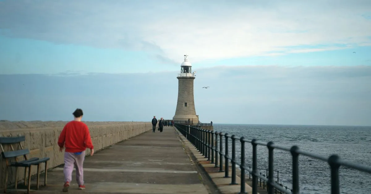 Coastal pier with a lighthouse and people walking during daytime, offering a serene seaside view.