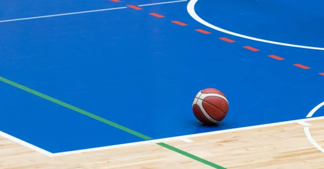 A basketball rests on a vibrant blue indoor court with wood flooring and colorful lines.