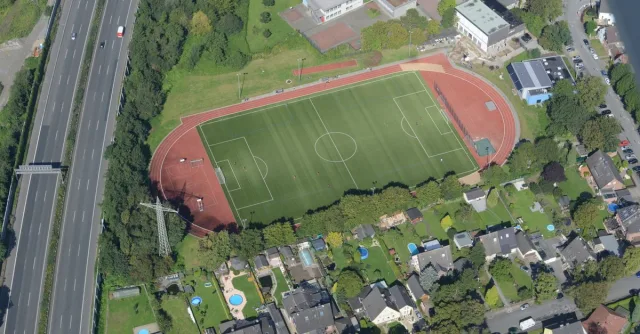 Aerial shot of a football field, roads, and houses amidst greenery, captured from above.